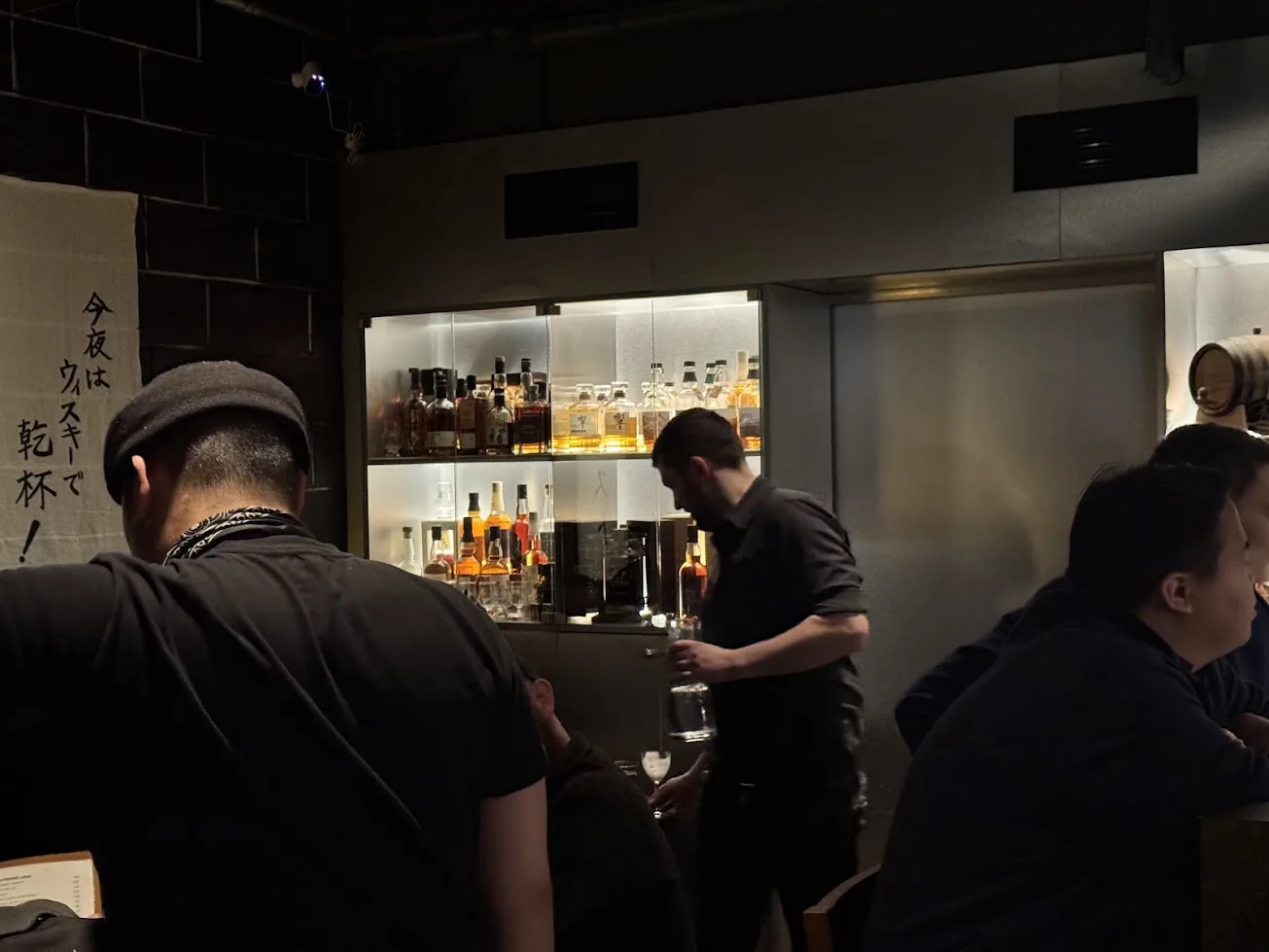 Bar Besuto at night: a bartender in front of a lit glass cabinet of Japanese whisky, with a calligraphy banner on the wall