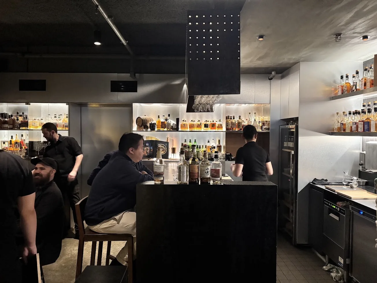 The Bar Besuto room: drinkers at a dark timber counter lined with bottles, and lit shelves of whisky behind