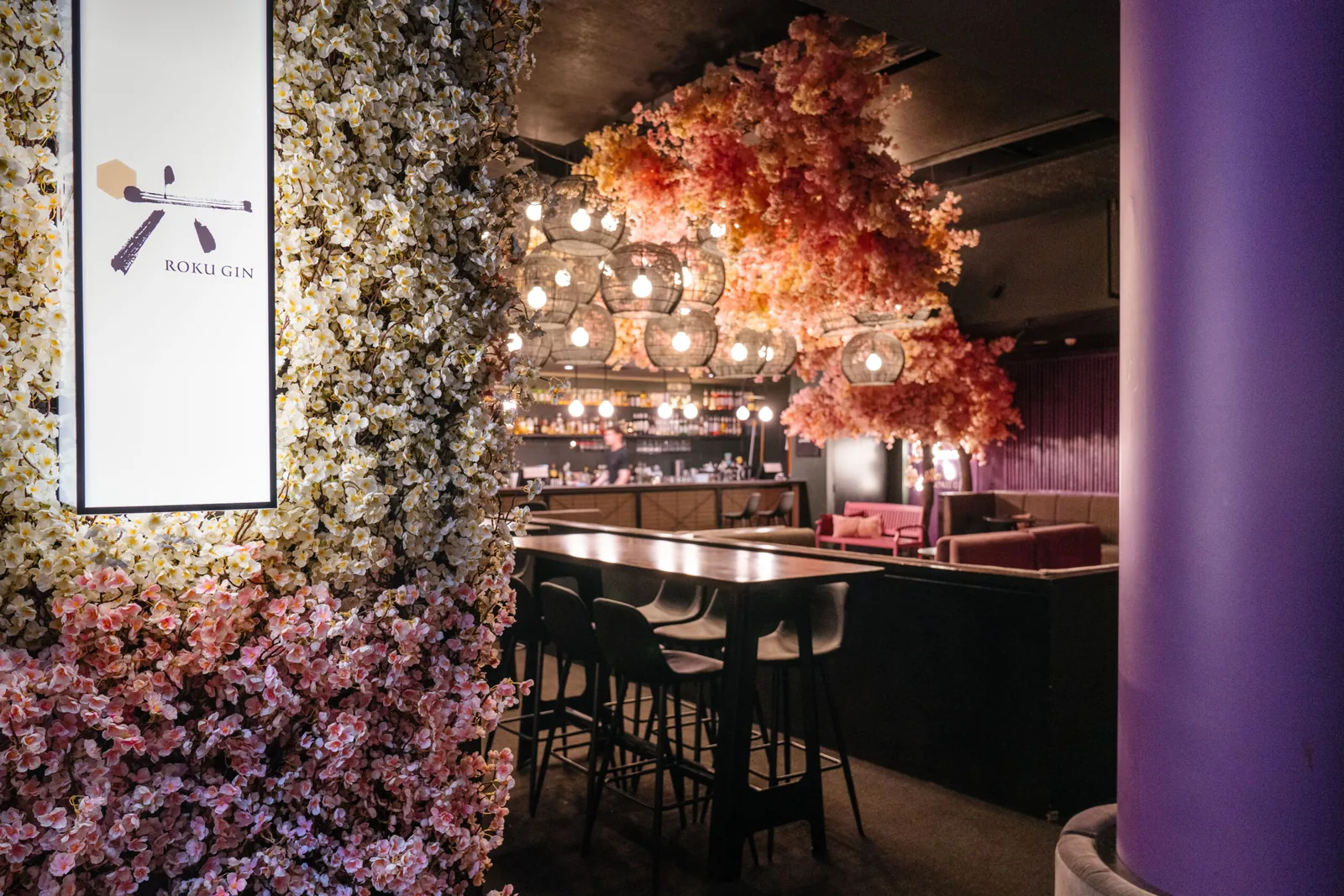 Inside Goody Two's: high tables and booths under a ceiling of pink cherry blossom and woven lanterns, with the lit back bar beyond