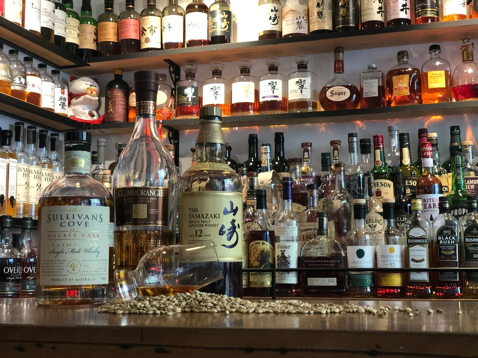 Whisky Den's back bar: shelves of whisky with Yamazaki 12 and Japanese-labelled bottles, and a nosing glass on its side among barley on the counter