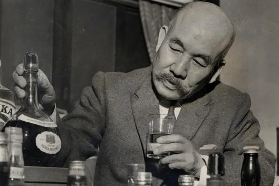Masataka Taketsuru in later life, seated at a table of Nikka bottles, pouring a glass.