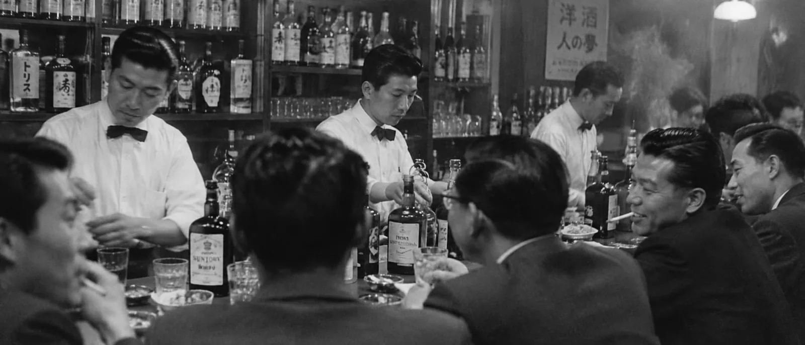 A crowded postwar Japanese bar. Bartenders in bow ties pour from bottles of Suntory and Torys whisky while men in suits drink and smoke along the counter.